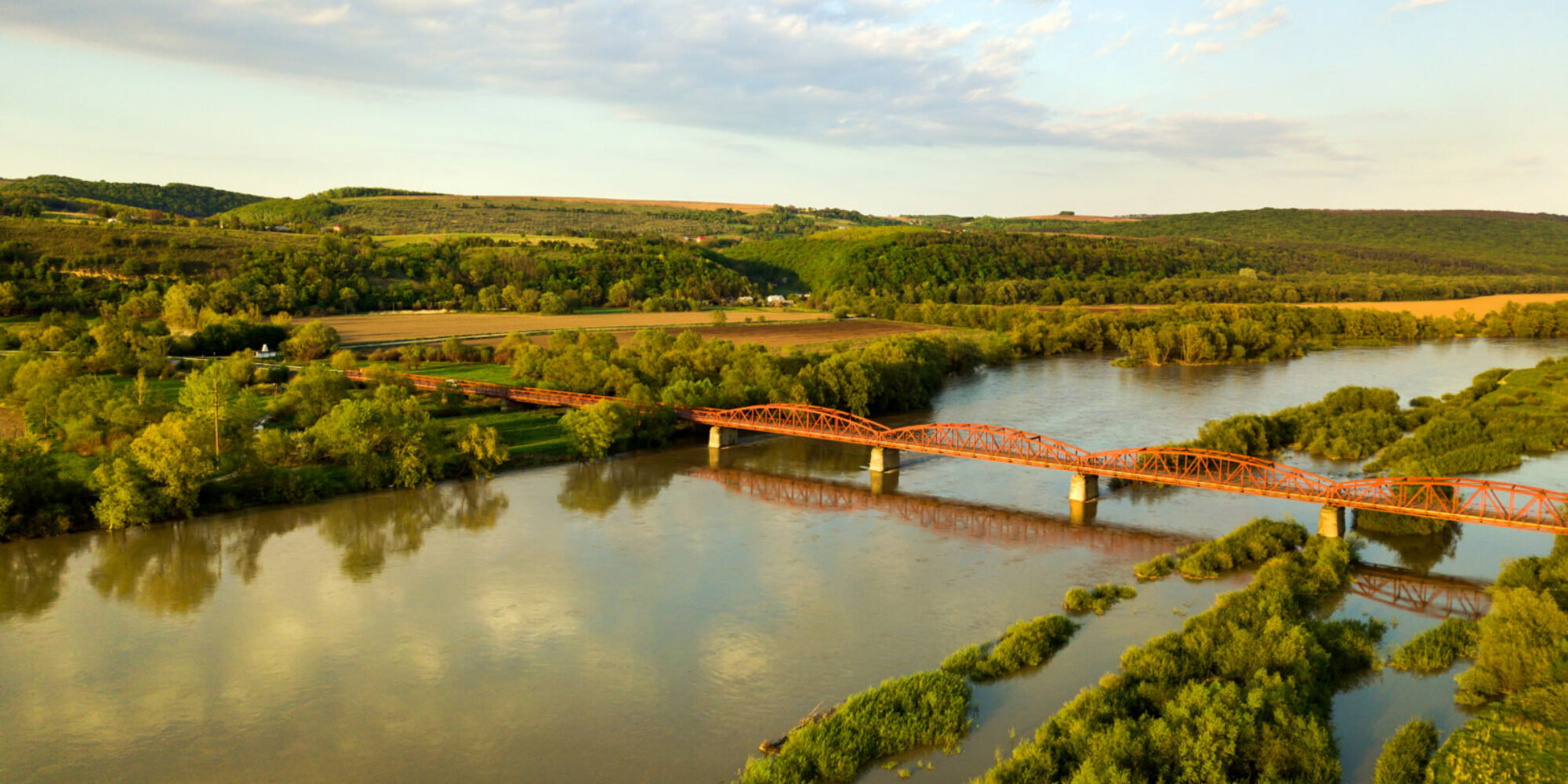 Aerial view of a narrow road bridge stretching over muddy wide river in green rural area.